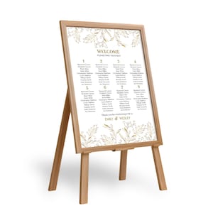 May include: A white wedding seating chart with gold floral accents and a wooden easel. The chart lists guest names and table numbers. The text at the top reads "Welcome, Please Find Your Seat". At the bottom, it says "Thank you for celebrating with us, Emily & Wesley".