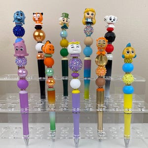 May include: A collection of decorative pens featuring character-themed toppers. Each pen has a unique design with colorful beads and a character figurine. The pens are displayed on a clear, tiered stand, showcasing their variety and detail.