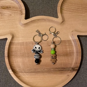 May include: Two keychains with character charms rest on a wooden Mickey Mouse-shaped tray. One keychain features a Jack Skellington charm with black and white beads. The other has a brown character charm with green and wooden beads. Both have silver key rings.