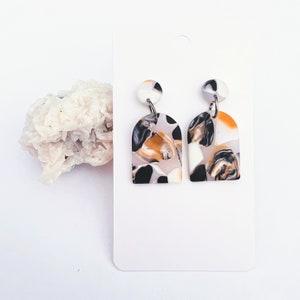 May include: A pair of white, black, and orange arch-shaped earrings with a marbled pattern. The earrings are made of polymer clay and have silver hooks.