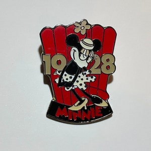 May include: A gold and black pin featuring Minnie Mouse in a red and white dress, standing in a red chair with the number 1928 on it. The text "MINNIE" is on the bottom of the pin.