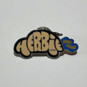 May include: A black and white enamel pin featuring a cartoon car with the word "HERBIE" in large letters. The car has a blue and yellow cloud coming out of the back with the text "RIDES AGAIN" and the year "1974" printed on the bottom.