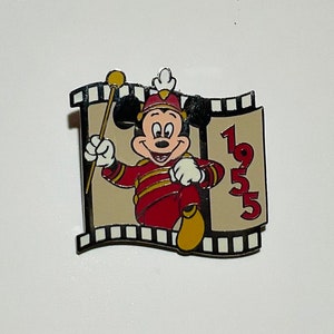 May include: A gold and red Mickey Mouse pin with a film strip border. Mickey is dressed as a bandleader and holding a baton. The year 1955 is printed on the film strip.