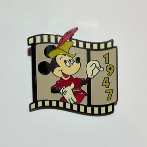 May include: A gold and black film strip pin featuring Mickey Mouse dressed as Robin Hood. The film strip has the year 1947 printed on it.