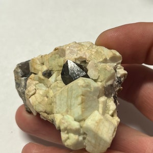 May include: A black, pointed crystal sits atop a cluster of white and green crystals. The crystals are embedded in a rough, grey rock.