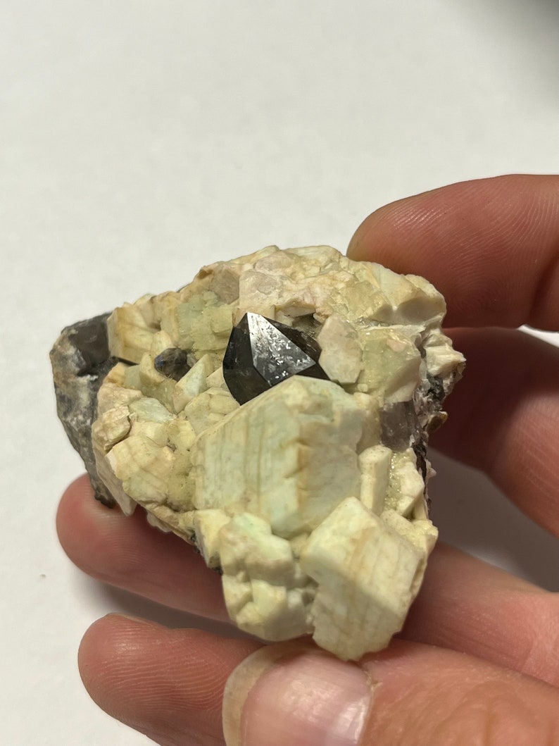 May include: A black, pointed crystal sits atop a cluster of white and green crystals. The crystals are embedded in a rough, grey rock.