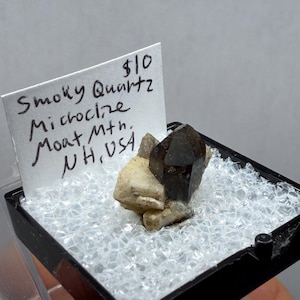 May include: A small, dark smoky quartz crystal specimen with a lighter-colored base, displayed in a clear plastic case. A handwritten label identifies it as "Smoky Quartz Microcline Moat Mtn. NH, USA."
