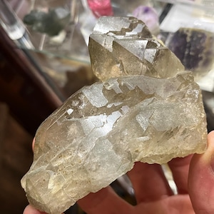 Puede incluir: Una mano sostiene un gran grupo de cristales translúcidos de color amarillo claro. El cristal tiene múltiples facetas y una superficie rugosa y texturizada. Otros cristales en tonos rosa, verde y morado son visibles en el fondo.