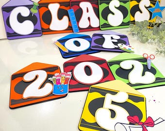 Könnte beinhalten: Abschlussbanner „Klasse von 2025“ mit bunten, kreidenförmigen Buchstaben und Zahlen. Die Buchstaben sind weiß mit schwarzen Umrandungen und die Zahlen sind weiß mit schwarzen Umrandungen. Das Banner ist mit Schulutensilien wie Bleistiften, Buntstiften, einer Schere, einem Lineal, einer Farbpalette und einer Abschluss-Kappe verziert.