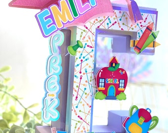 May include: A purple letter E decorated with school-themed items, including a red apple with a schoolhouse, a blue backpack, a yellow wagon with the numbers 123, a yellow wagon with the letters ABC, a ruler, pencils, crayons, and a push pin. The letter E is decorated with a colorful pattern of lines and dots. The letter E is topped with a pink graduation cap with the name "Emily" written on it in colorful letters.