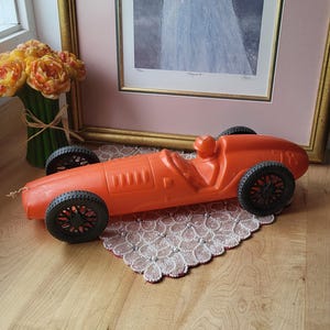 Peut inclure: Une voiture de course jouet orange vif avec des roues noires et une figurine de pilote. Le jouet en plastique est posé sur un napperon en dentelle décoratif. Une image encadrée et des fleurs sont en arrière-plan.