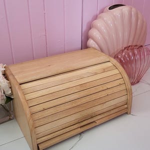May include: A wooden bread bin with a curved, slatted lid. The rectangular box has a pull-out drawer. The background features a pink wall and decorative vases, suggesting a kitchen or dining room setting.