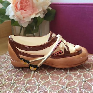 Op de afbeelding: Een keramische schoenvormige plantenbak in bruin- en crèmetinten, met decoratieve details. De plantenbak heeft een paar gekruiste golfclubs en staat tegen een achtergrond van een roze kralen oppervlak en bloemen.