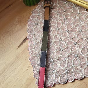 Op de afbeelding: Een veelkleurige leren riem met een messing gesp. De riem is gemaakt van aan elkaar gestikte delen van bruin, groen, marineblauw, rood en zwart leer. De riem is ongeveer 2,5 cm breed.