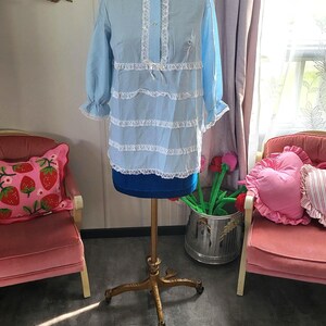 May include: A light blue vintage-style blouse with white lace trim and a matching pair of bloomers. The blouse is displayed on a mannequin, showcasing its long sleeves and ruffled details. The bloomers are on the floor.