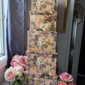 Könnte beinhalten: Ein Stapel von sechs dekorativen Boxen mit einem Vintage-Weihnachtsmotiv. Die Boxen sind aus Pappe und haben ein rosa-cremefarbenes Farbschema. Jede Box zeigt eine andere Collage von Vintage-Weihnachtsbildern, darunter Engel, Weihnachtsmann und Kinder. Die Boxen sind übereinander gestapelt und bilden so ein festliches Display.