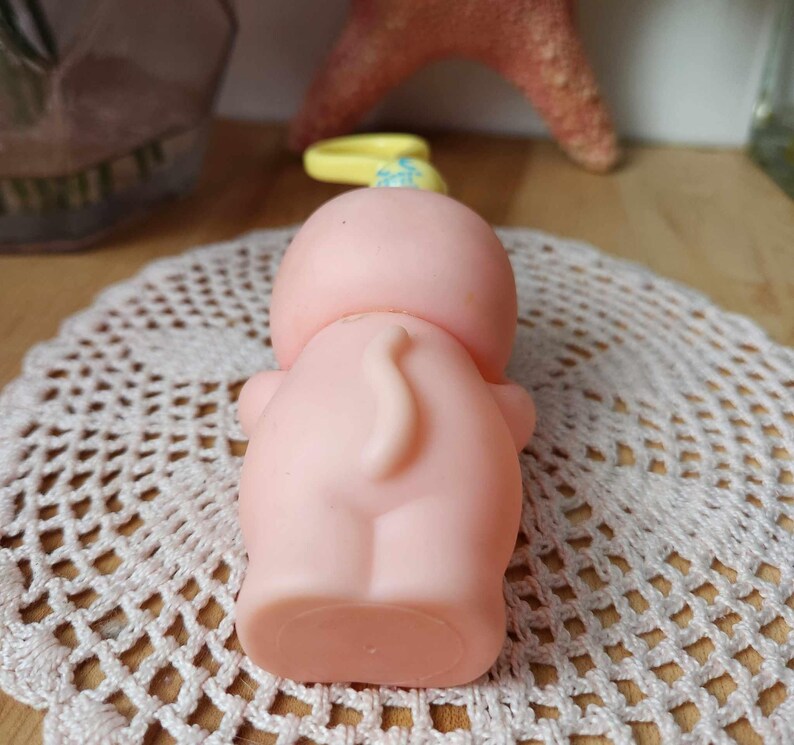 Puede incluir: Un juguete de goma rosa con una cola amarilla y una cabeza azul y amarilla. El juguete est&aacute; sentado en un encaje blanco de ganchillo.