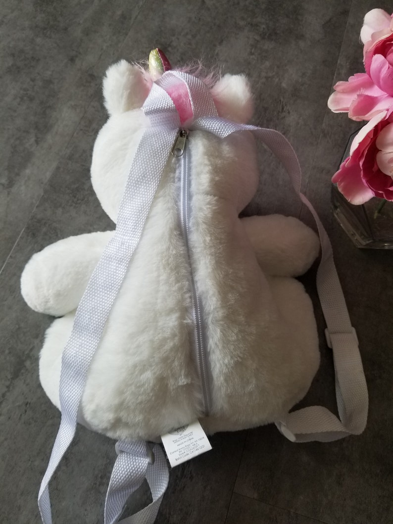 May include: White plush unicorn backpack with a pink and gold horn. The backpack has a zipper closure and white straps.