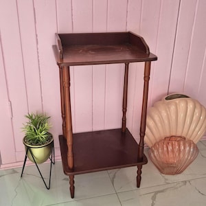 Puede incluir: Una mesa auxiliar de madera de color marrón oscuro con dos niveles, cuatro patas torneadas y un pequeño borde trasero en el estante superior. La mesa cuenta con una superficie rectangular y un estante inferior a juego para dos niveles.