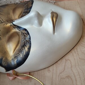 May include: A white and gold masquerade mask with black painted details. The mask has a stylized eye and mouth design.