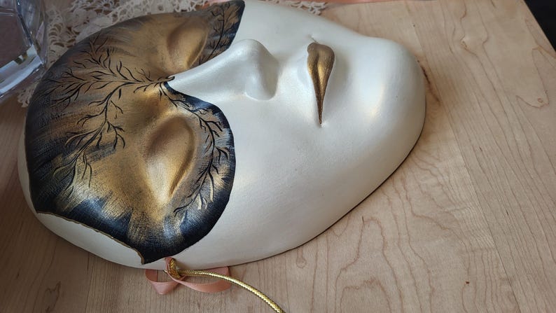May include: A white and gold masquerade mask with black painted details. The mask has a stylized eye and mouth design.