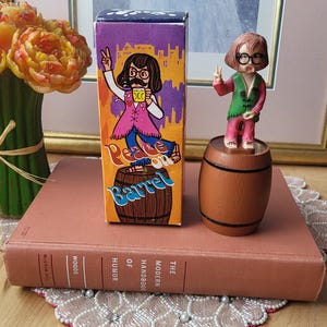 Pode incluir: Um item de novidade vintage com uma pequena estatueta em cima de um barril em miniatura, ao lado de uma caixa com as palavras "Peace on Barrel". A caixa e a estatueta retratam um personagem de desenho animado com óculos e um sinal de paz. Os itens estão em um livro.