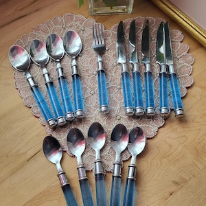 Puede incluir: Un juego de cubiertos de acero inoxidable con mangos de acrílico azul. El juego incluye cucharas, tenedores y cuchillos, dispuestos sobre un salvamanteles decorativo. Los mangos son transparentes y las partes metálicas son plateadas.