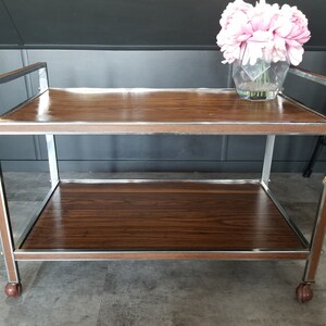 May include: A two-tiered serving cart with a brown wood top and chrome frame. The cart has wheels and is perfect for serving drinks and snacks.