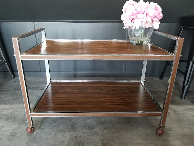 May include: A two-tiered serving cart with a brown wood top and chrome frame. The cart has wheels and is perfect for serving drinks and snacks.