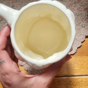May include: A white ceramic mug with a handle, viewed from above. The interior of the mug has a light yellow hue. The mug is held by a person, with a wooden surface and decorative beaded placemat in the background.