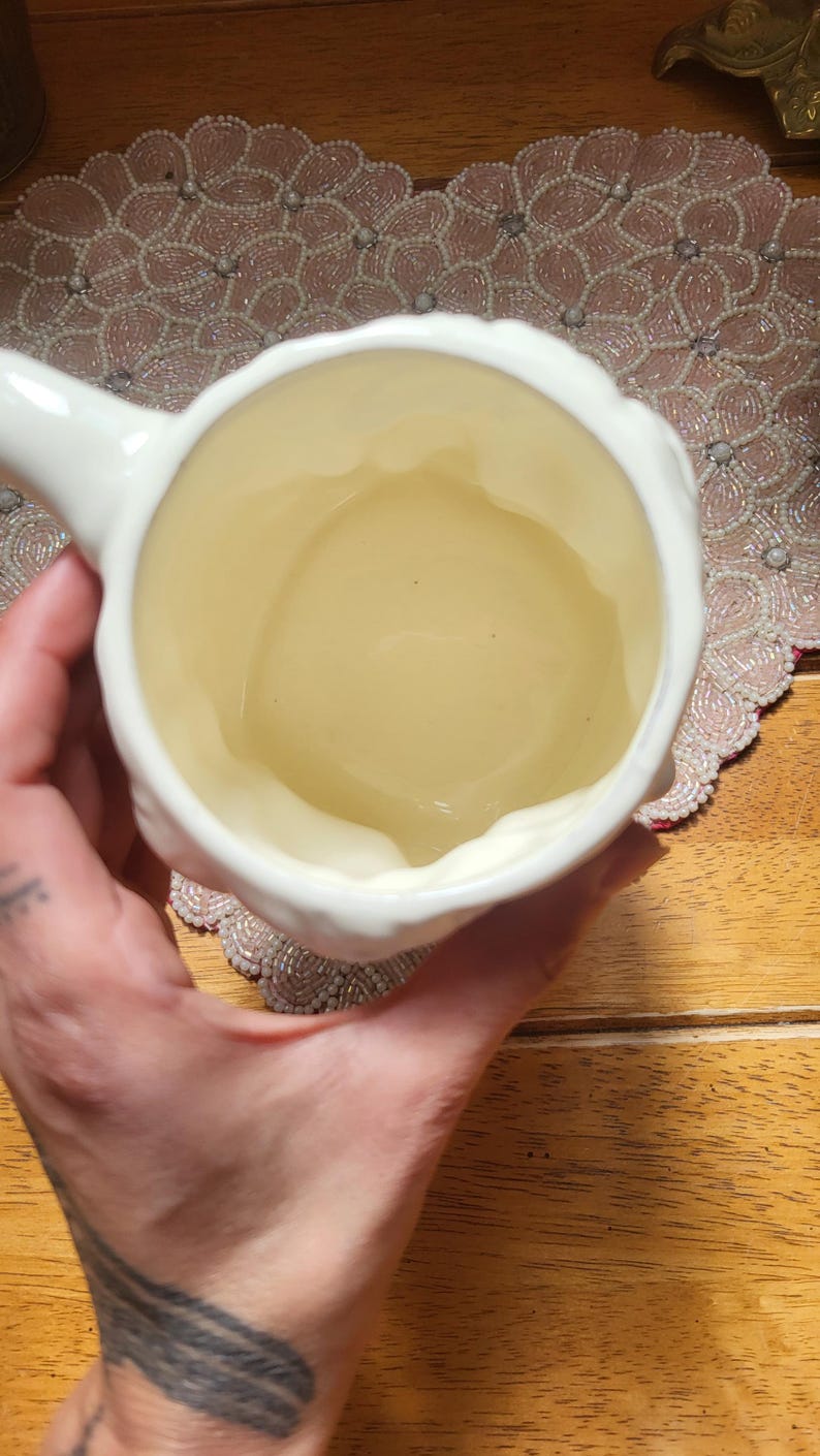 May include: A white ceramic mug with a handle, viewed from above. The interior of the mug has a light yellow hue. The mug is held by a person, with a wooden surface and decorative beaded placemat in the background.