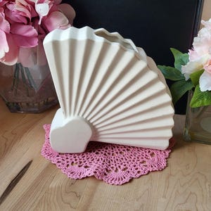 Könnte beinhalten: Eine cremefarbene, fächerförmige Vase mit gewellter Oberseite, die auf einer rosa gehäkelten Tischdecke steht. Die Vase steht auf einer Holzoberfläche, mit rosa Blumen im Hintergrund.
