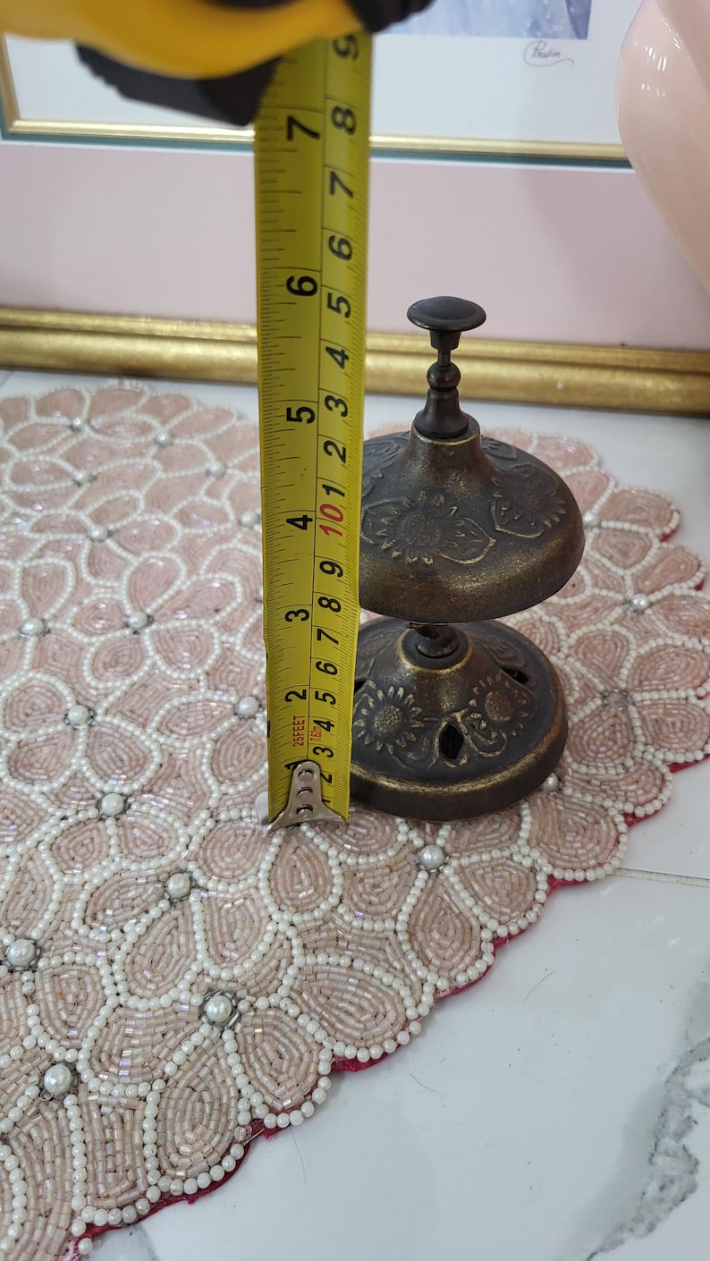 Puede incluir: Una campana de lat&oacute;n antiguo, de aproximadamente 7,6 cm de altura, con un patr&oacute;n floral decorativo. La campana se mide con una cinta m&eacute;trica amarilla. La campana se encuentra sobre un mantel individual rosa con cuentas y un dise&ntilde;o floral.