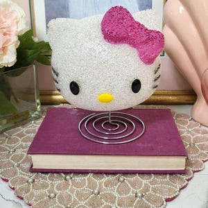 May include: A decorative Hello Kitty head with a pink bow, black eyes, and a yellow nose, sits atop a purple book. The head is made of a clear, textured material. A silver wire book stand is under the book. The items are on a beaded placemat.