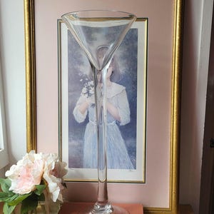 May include: A tall, clear glass martini glass with a long stem. The glass sits on a brown book, with a framed picture in the background. A vase of pink flowers is to the left.