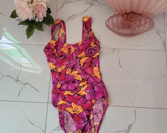 Pode incluir: Um fato de banho de uma peça com um padrão floral vibrante em tons de rosa, laranja e amarelo. O fato de banho tem um decote quadrado e alças largas. O fundo inclui uma parede rosa, flores e itens decorativos.