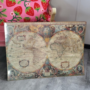 Puede incluir: Un mapa del mundo antiguo enmarcado con cartografía detallada, que presenta dos hemisferios e ilustraciones decorativas. El mapa se muestra sobre un suelo gris, con un cojín rosa con estampado de fresas en el fondo.