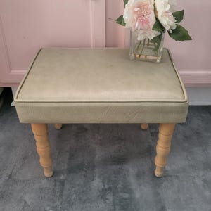 May include: A small, upholstered bench with a light beige cushion and turned wooden legs. The bench is topped with a clear glass vase holding a bouquet of pink and white flowers. The background features pink cabinets.