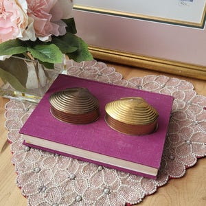 May include: Two seashell-shaped trinket boxes in bronze and gold tones rest on a closed, purple book. The boxes have a ribbed design. The book sits on a beaded, pink lace doily. A framed print and pink flowers are in the background.