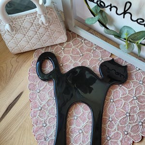 Peut inclure: Un objet décoratif noir brillant en forme de chat. Le chat a un design épuré et minimaliste avec une tête arrondie et un long corps arqué. Il est placé sur un napperon rose perlé. Un vase blanc en céramique en forme de sac à main est en arrière-plan.