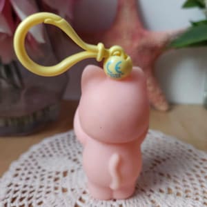 Puede incluir: Un llavero de goma rosa en forma de cerdo con un clip amarillo y una marca CE azul. El cerdo est&aacute; de pie sobre sus patas traseras con la cola enroscada.