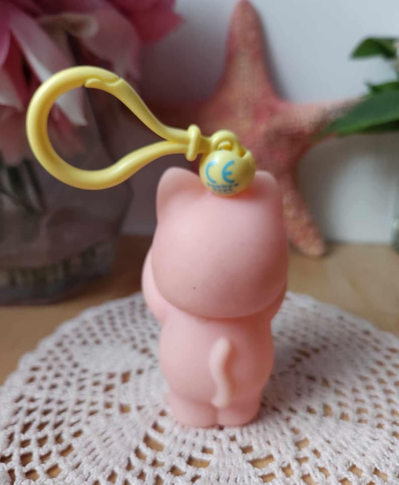 Puede incluir: Un llavero de goma rosa en forma de cerdo con un clip amarillo y una marca CE azul. El cerdo est&aacute; de pie sobre sus patas traseras con la cola enroscada.