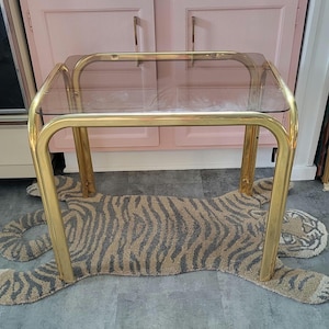 May include: A vintage side table with a clear glass top and a gold-colored metal frame. The table is placed on a rug shaped like a tiger, with a gray and tan striped pattern. The background features pink cabinets.