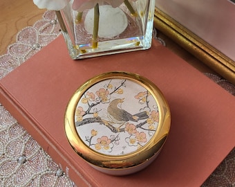 Könnte beinhalten: Eine runde, antike Schmuckdose mit goldenem Rand und Deckel mit einem Vogel auf einem blühenden Zweig. Die Dose liegt auf einem geschlossenen, rotbraunen Buch, mit einem Perlen-Platzset und einer Blumenvase im Hintergrund.
