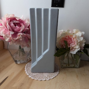 Può includere: Un vaso alto in ceramica grigia con linee verticali e un design geometrico. Il vaso è appoggiato su un sottopentola in pizzo bianco su una superficie in legno.