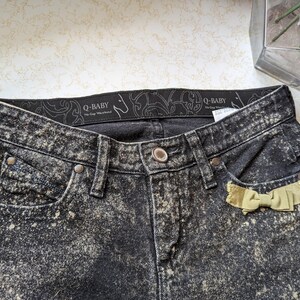 May include: Black acid wash denim jeans with a yellow bow on the back pocket. The jeans have a black waistband with the text "Q-BABY" in white.