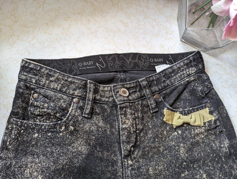 May include: Black acid wash denim jeans with a yellow bow on the back pocket. The jeans have a black waistband with the text "Q-BABY" in white.