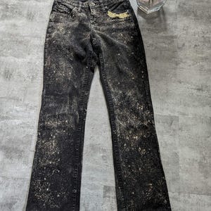 May include: Black denim jeans with a bleach splatter design. The jeans have a bootcut leg style.