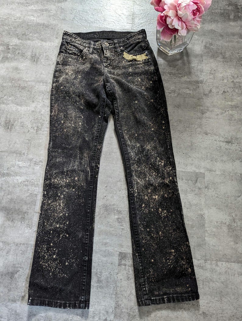 May include: Black denim jeans with a bleach splatter design. The jeans have a bootcut leg style.
