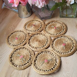 May include: Set of eight woven straw placemats with pink flower accents. The placemats are arranged in a circular pattern on a wooden table.